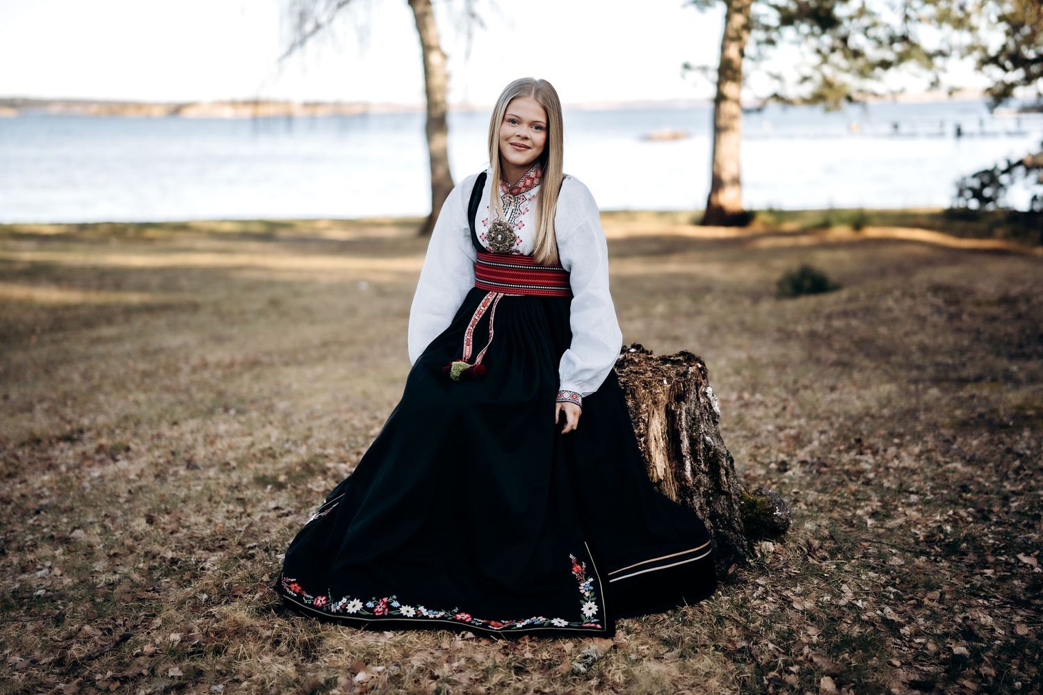Konfirmasjonsfotografering - Ei tenåringsjente som sitter på en stubbe og smiler mot kamera, i sin beltestakkbunad.