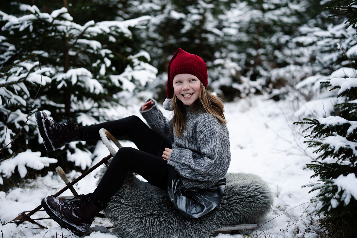 Julefoto av en jente med nisselue som. sitter på en kjelke i skogen dekket av snø.