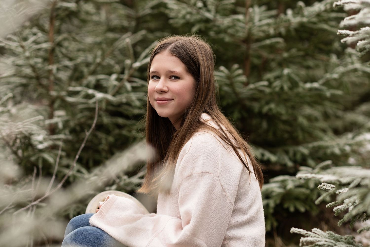 Barnefotografering av ei eldre jente som sitter i en furuskog med lys genser. Smiler mot kamera.
