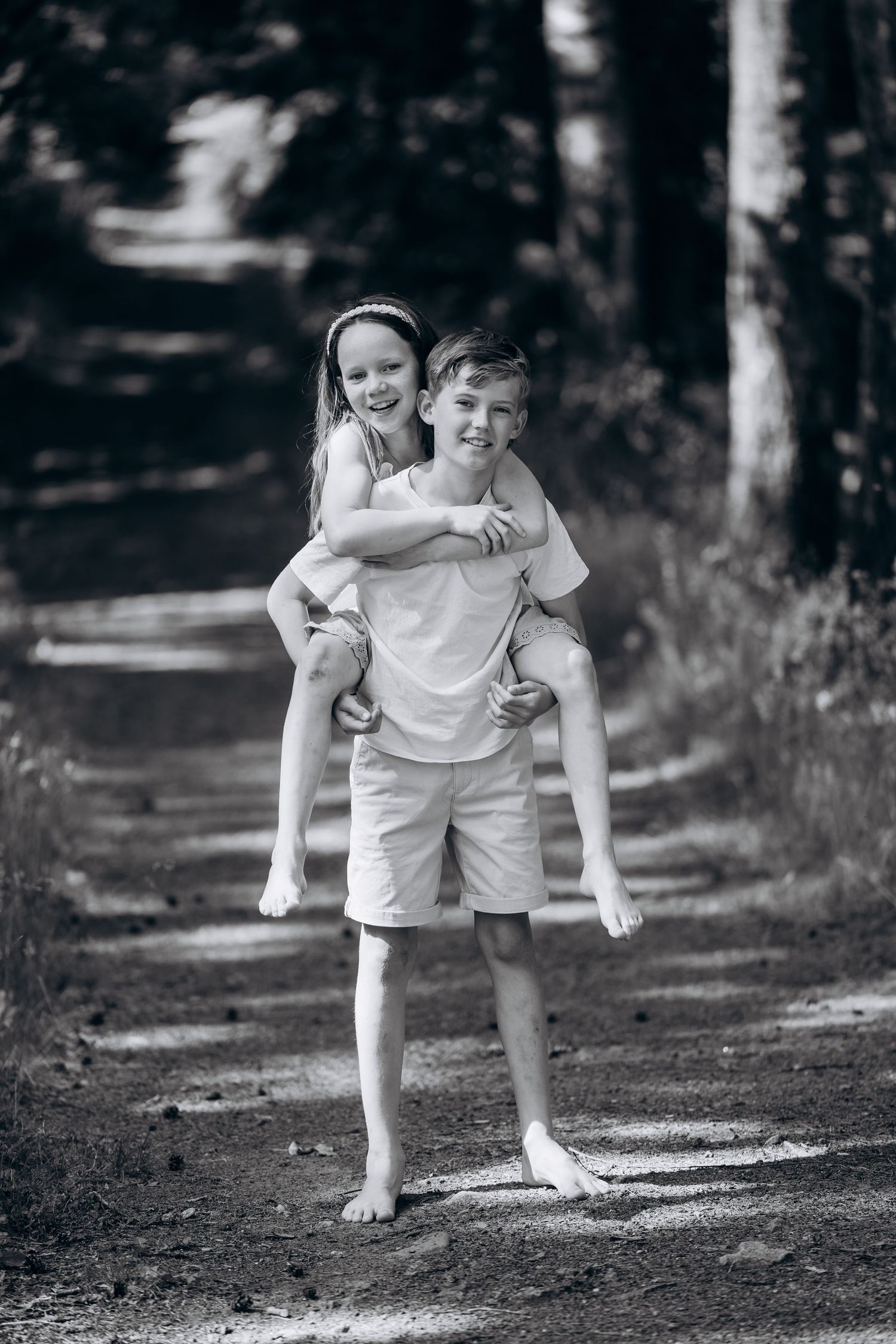 Familiefotografering i svart-hvitt av to barn på en skogssti, der det ene bærer det andre på ryggen.
