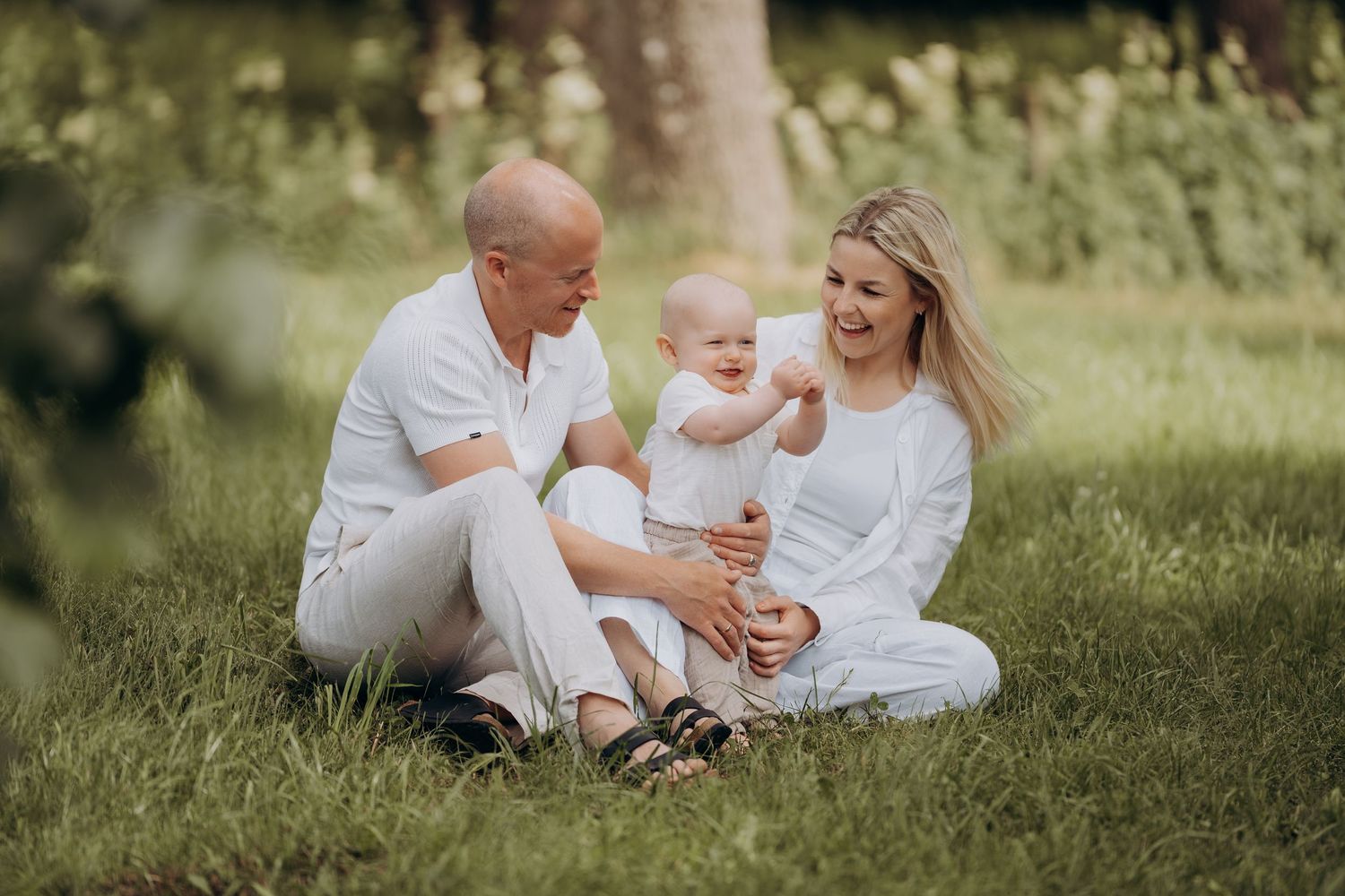 Familieportrett av mor, far og 1 åring som sitter i gresset en varm sommerdag.