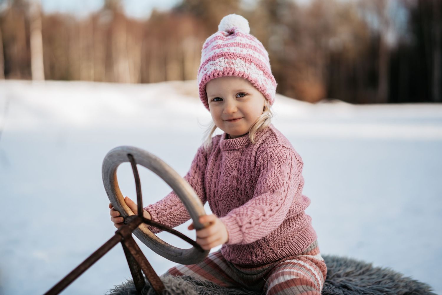 Et barnefoto av ei 4 år gammel jente som sitter på en kjelke i rosa strikkede klær. Hun smiler mens hun ser inn i kameraet.