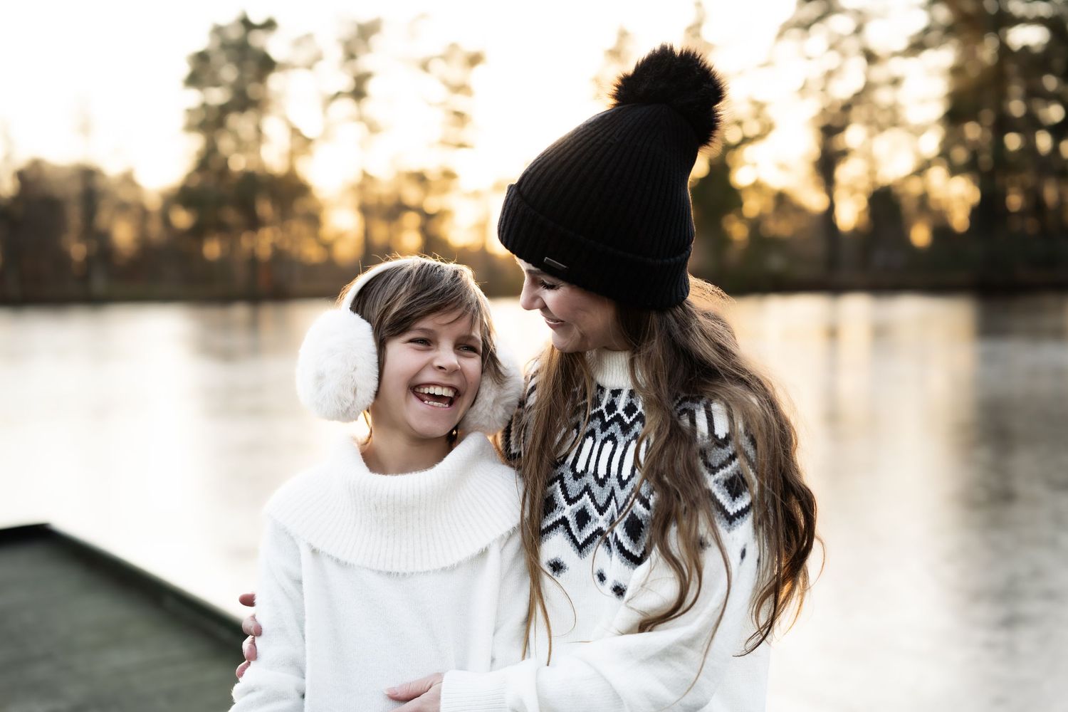 Mor og datter fotografering. Mor og datter står ute om vinteren i solnedgangen foran en innsjø. Begge har lyse klær og smiler til hverandre.