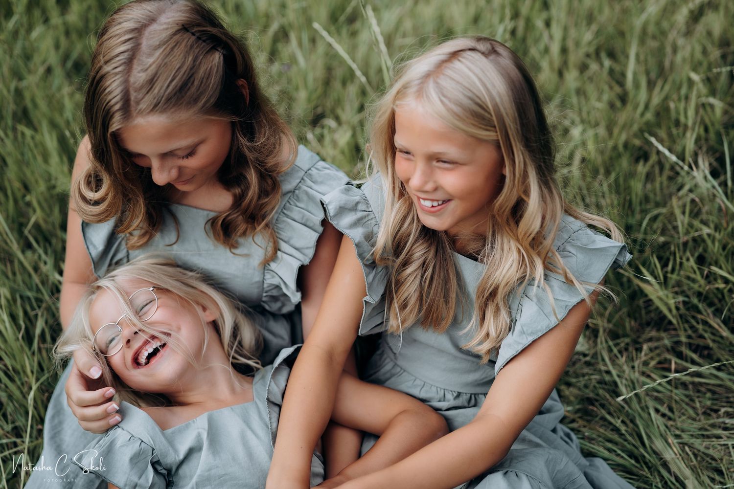 Søskenfotografering - Tre søstre sitter i kjole nede i gresset i enga og ler og smiler mot hverandre.
