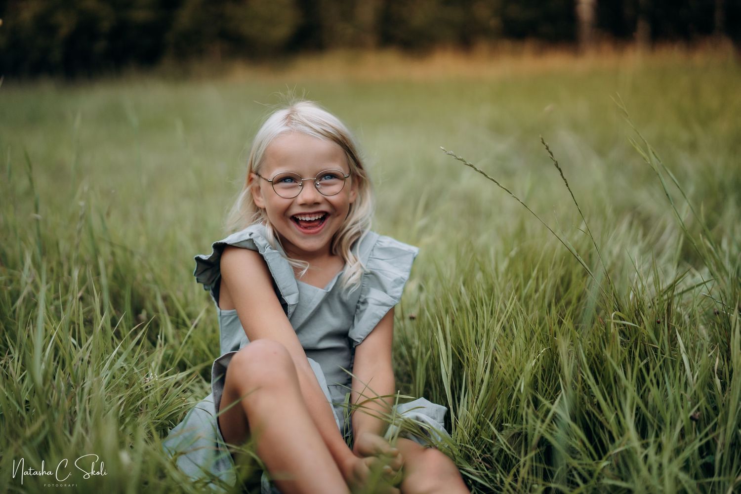 Barnefotografi - Ei smilende jente i kjole som sitter i en eng, en varm sommerkveld.