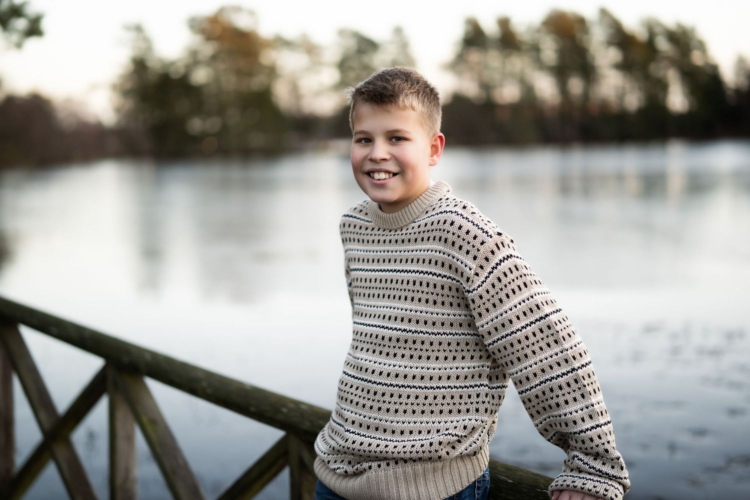 Fotografering av en tenåringsgutt i strikkegenser som står ute foran en innsjø om vinteren og smiler til kamra.