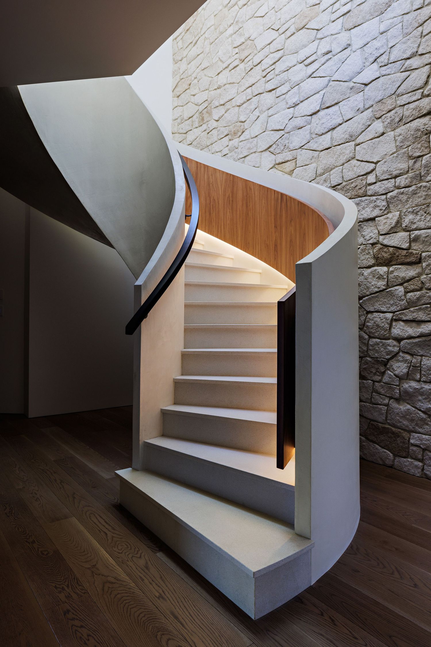 Elegant curved staircase with white steps, wooden accents and textured stone wall in modern interior design.