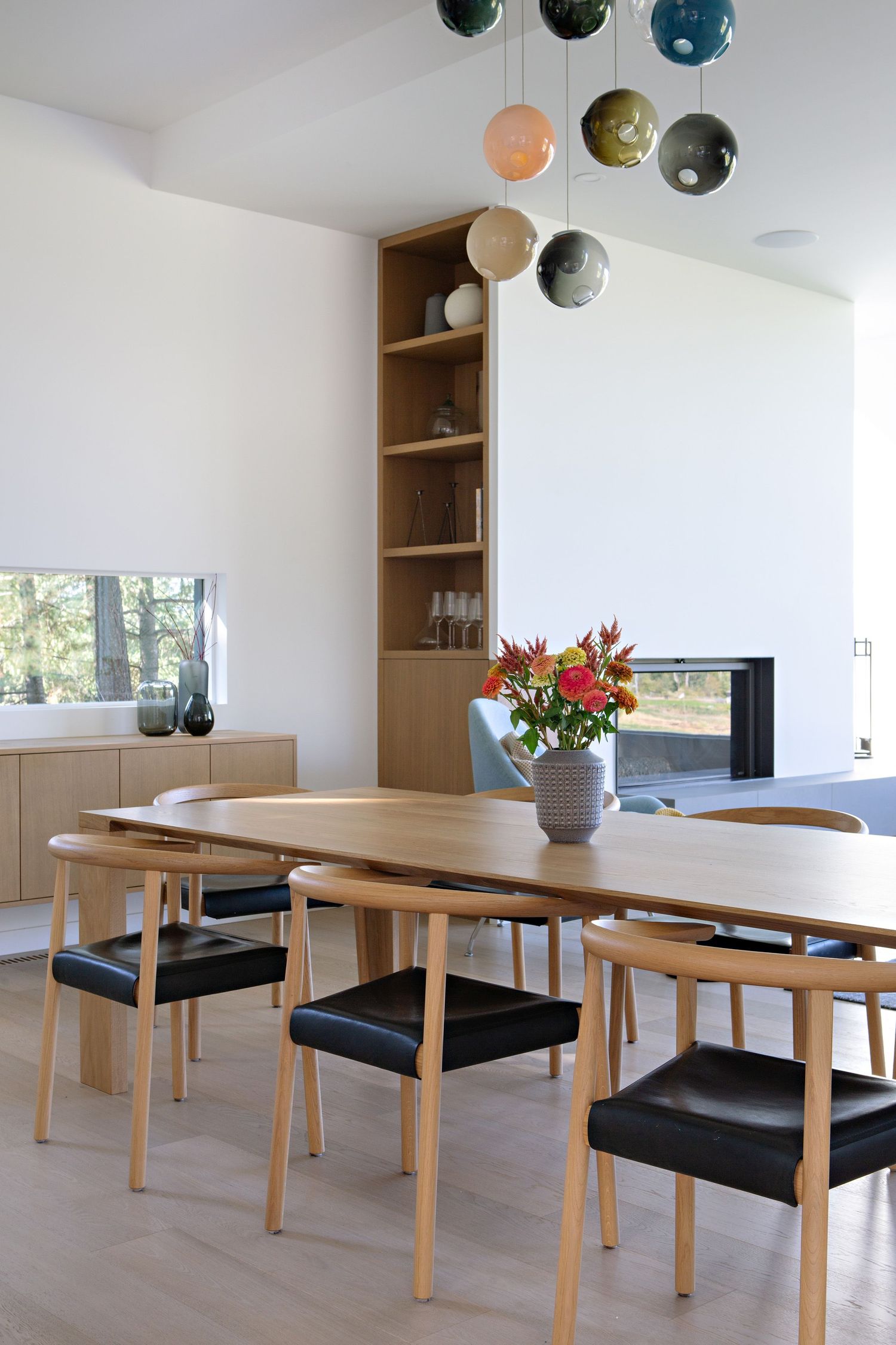 Modern dining room with wooden table, black leather chairs, floating orb pendant lights and built-in shelving by a window.