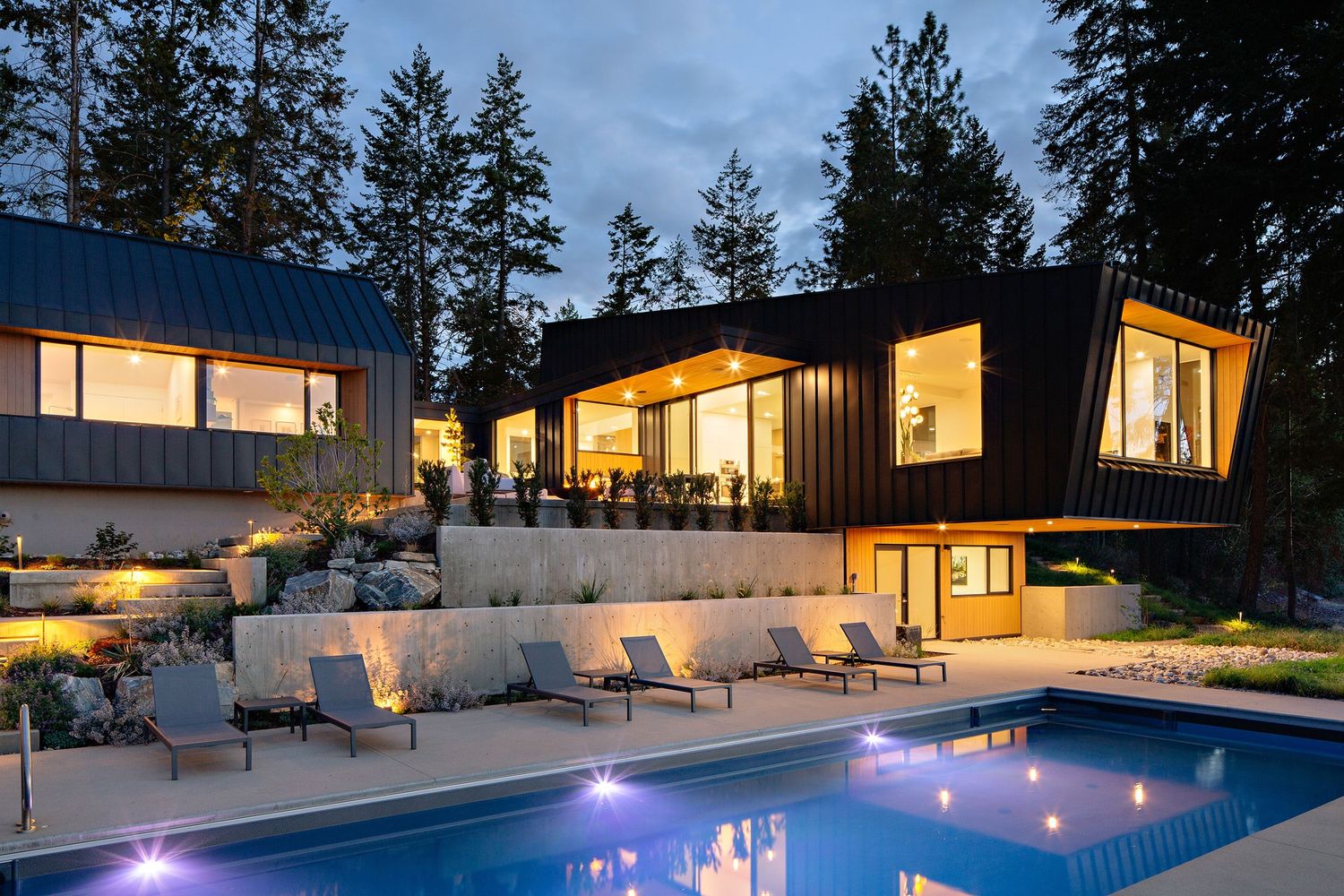 Luxurious modern home with glowing interior lights, geometric architecture and illuminated swimming pool at twilight.