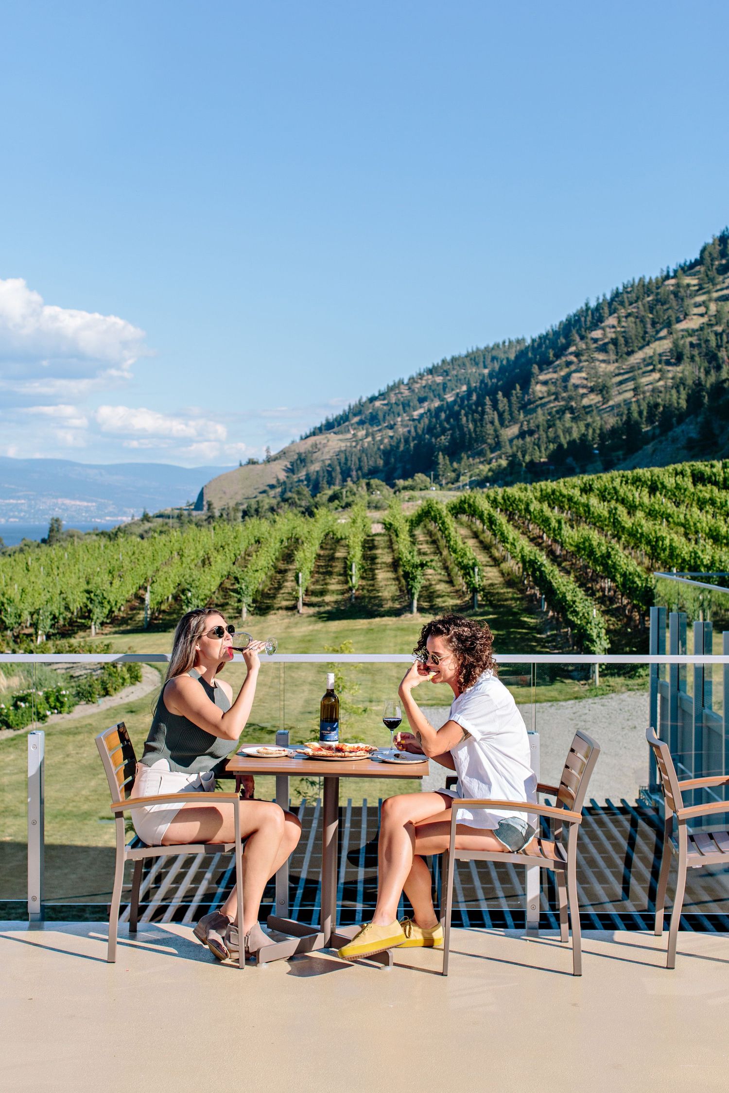Two people enjoy wine and snacks on an outdoor patio overlooking vineyard rows on a sunny mountain hillside.