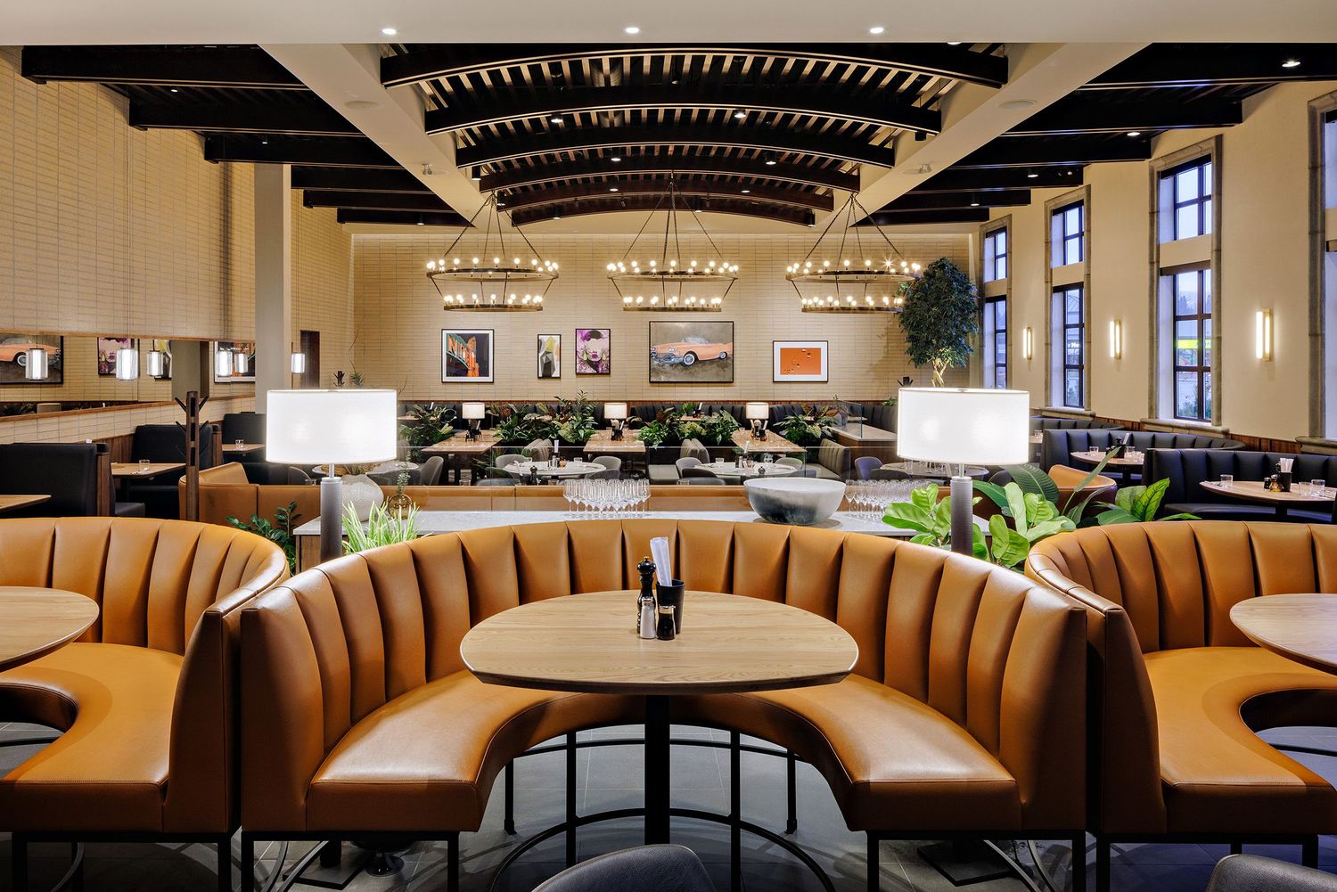 Elegant restaurant interior with curved brown leather booths, round tables, and grand chandeliers hanging from geometric ceiling.