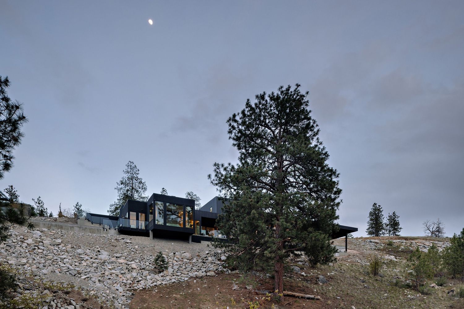 A modern cabin sits perched atop a rocky hillside with pine trees under a moody evening sky.