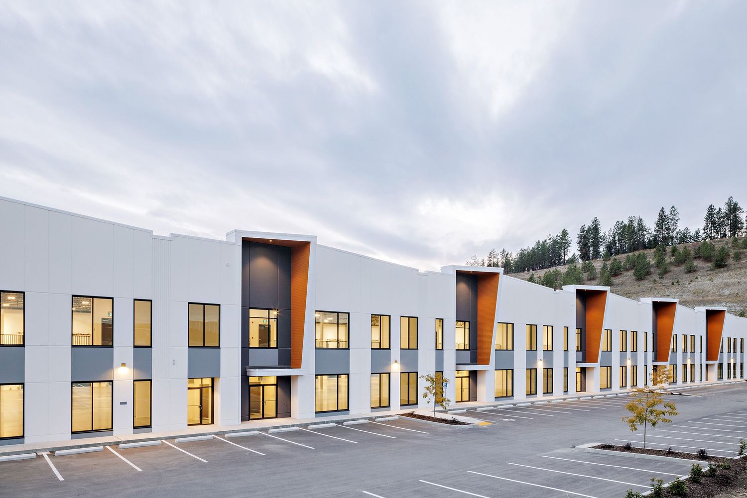 Modern industrial building with alternating gray and orange facade panels and illuminated windows at dusk.