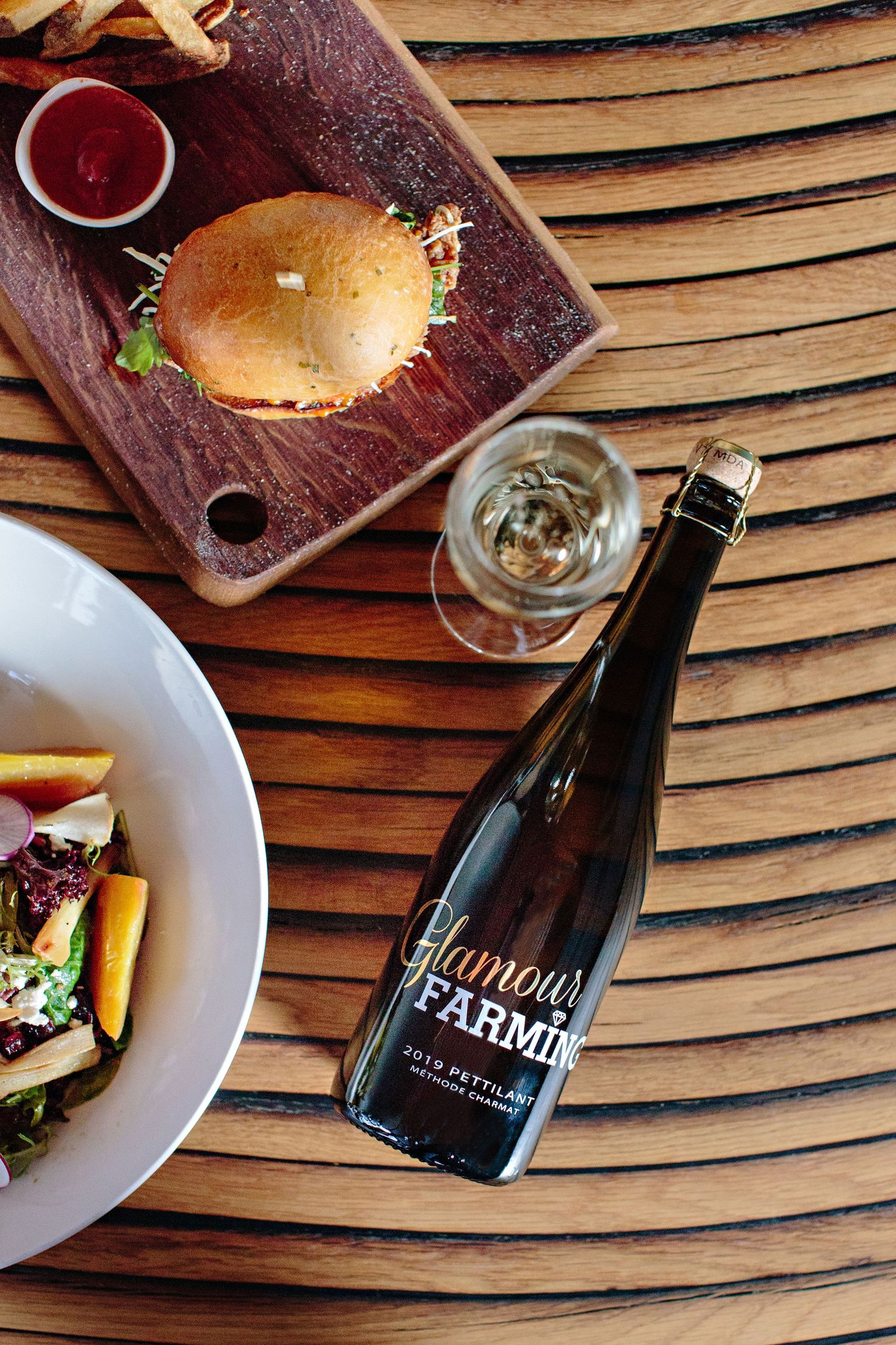 Gourmet burger and salad served with a bottle of Parmin wine on a wooden table.