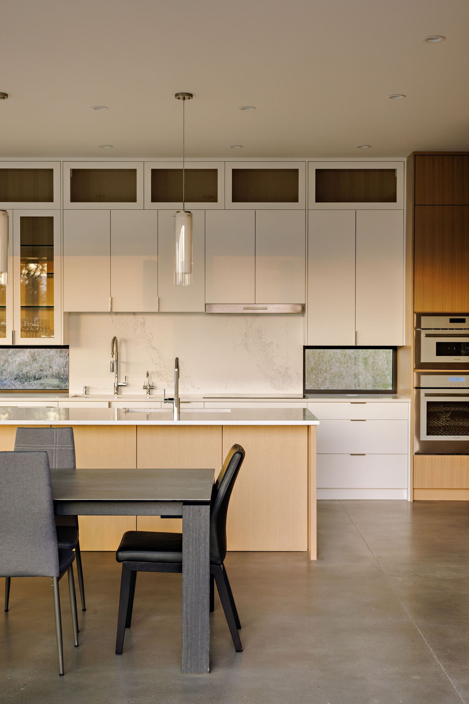 Modern kitchen interior with white cabinets, pendant lights, wooden accents and gray dining chairs on concrete flooring.