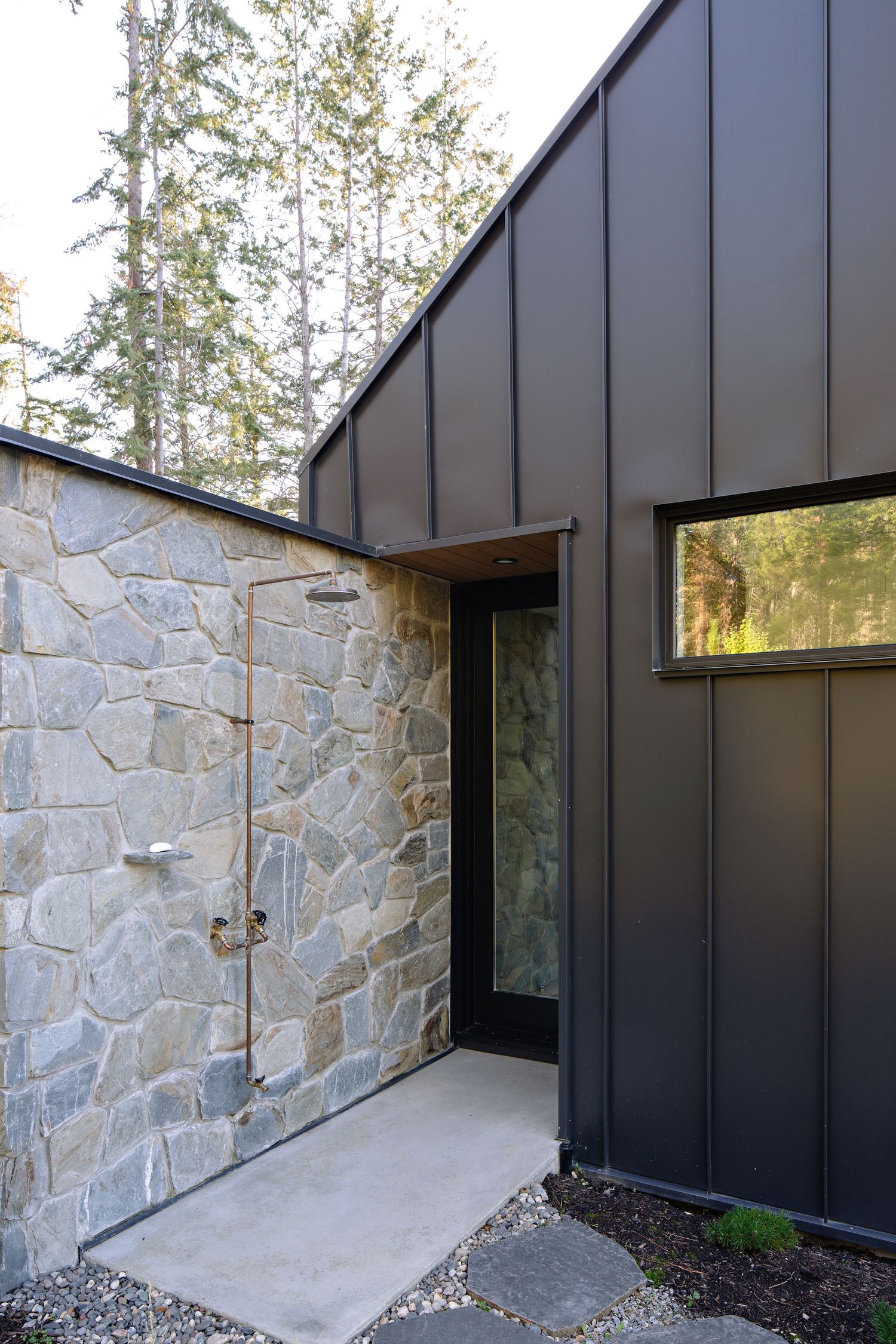 Modern home exterior featuring contrasting stone wall and black metal siding with floor-to-ceiling windows and concrete pathway.