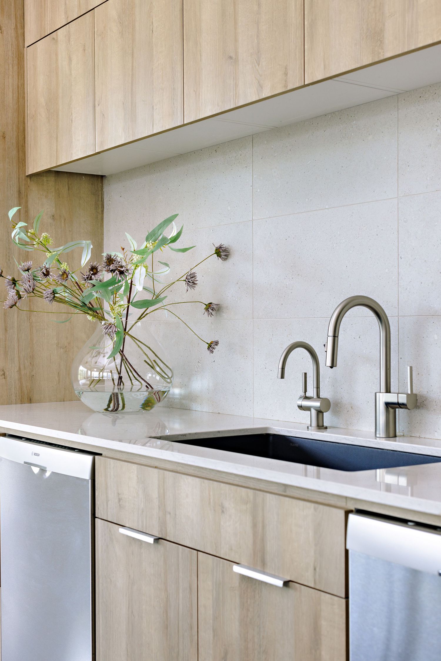 Modern kitchen with light wood cabinets, white counters, black sink, dual faucets and a glass vase with greenery.