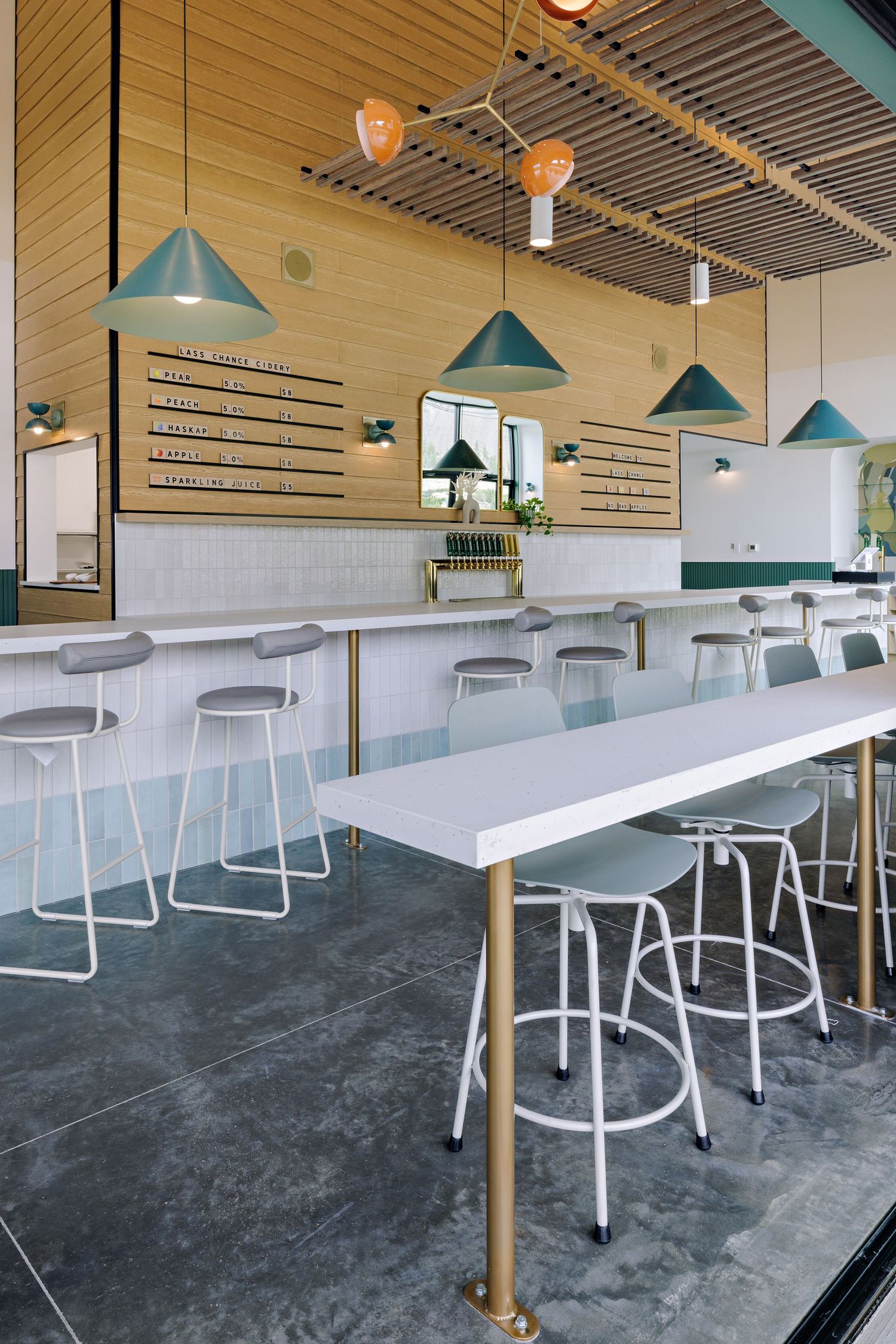 Contemporary dining space with turquoise pendant lights, white stools, and long communal tables on concrete floors.