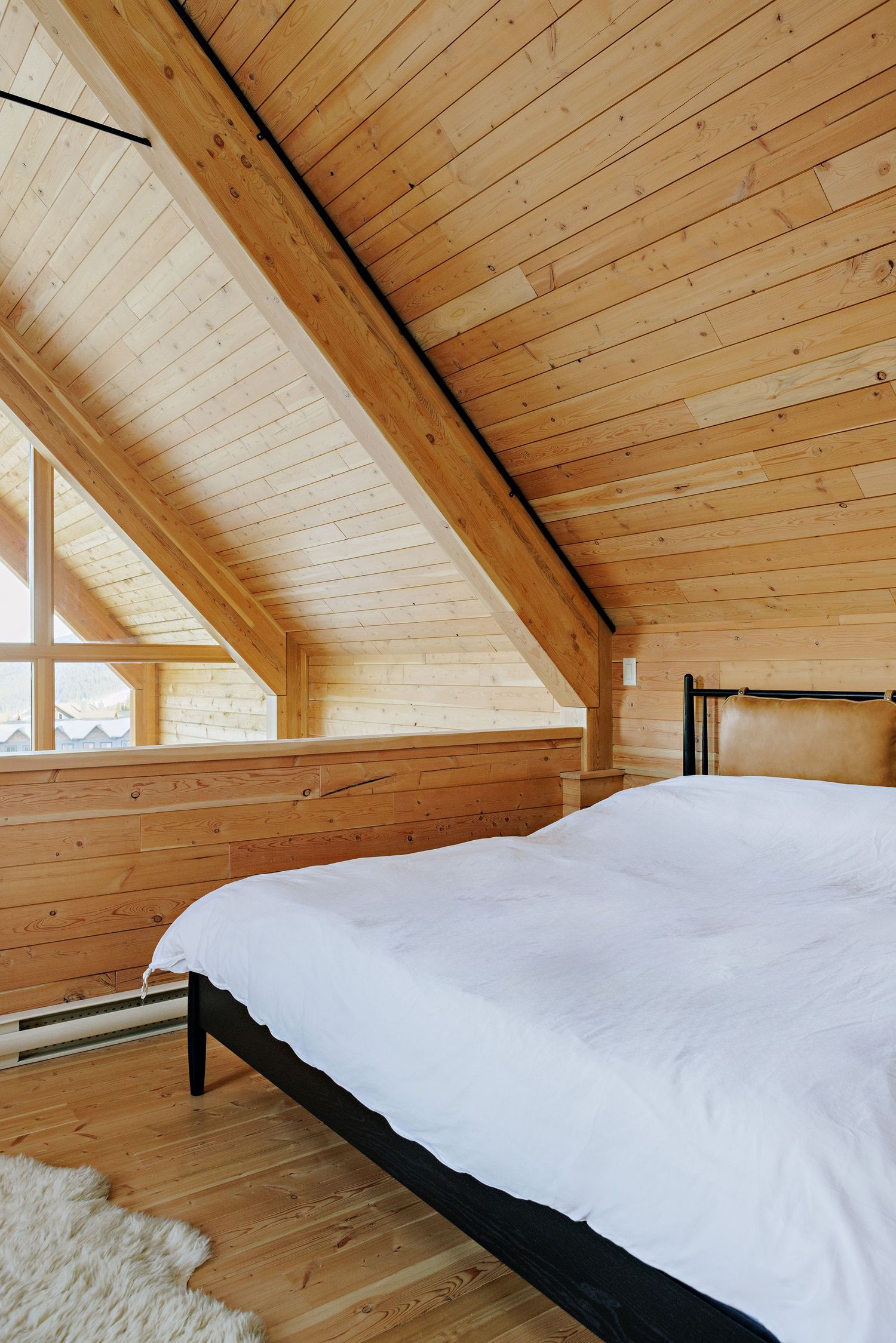A cozy bedroom with wooden walls and ceiling features a black metal bed frame with white bedding and a triangle window.