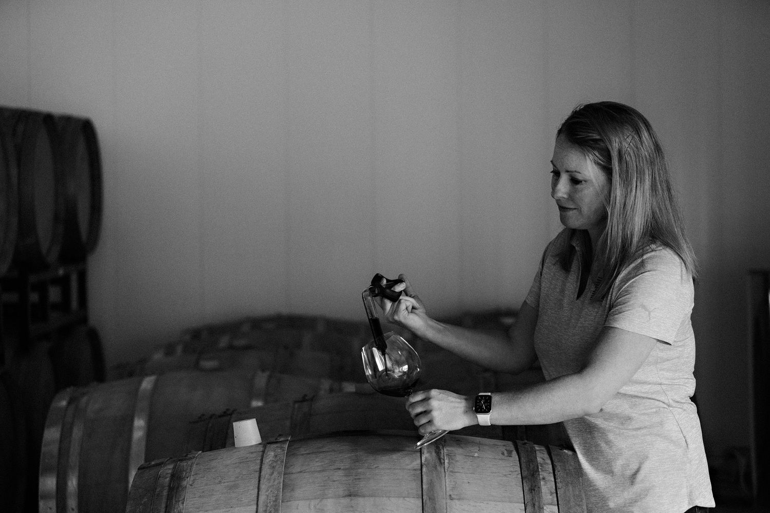 A person examines and works with wine barrels in a dimly lit cellar setting captured in black and white.