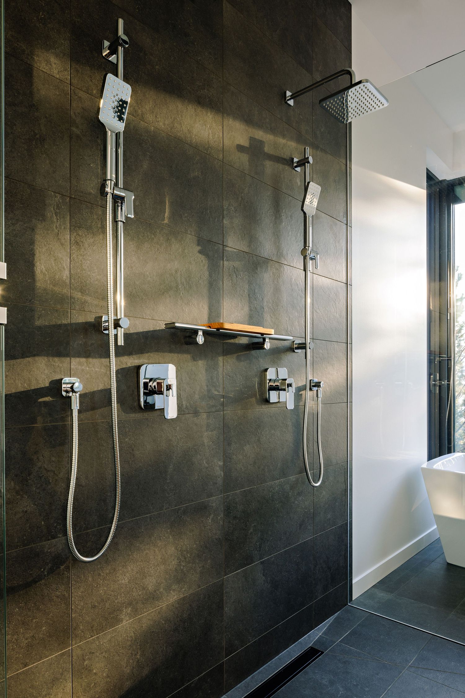 Modern walk-in shower with dark grey tile walls, dual showerheads, and chrome fixtures mounted on a textured wall.