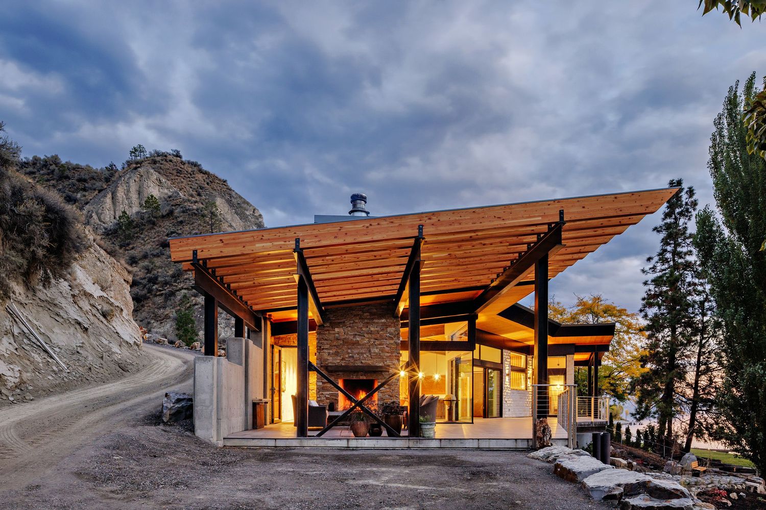 Modern mountain home with angular wooden roof and large windows glows warmly at dusk against rocky landscape.