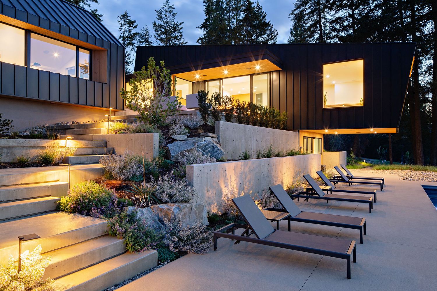 Modern tiered patio with chaise lounges and landscaped gardens illuminated at dusk beside contemporary luxury home.