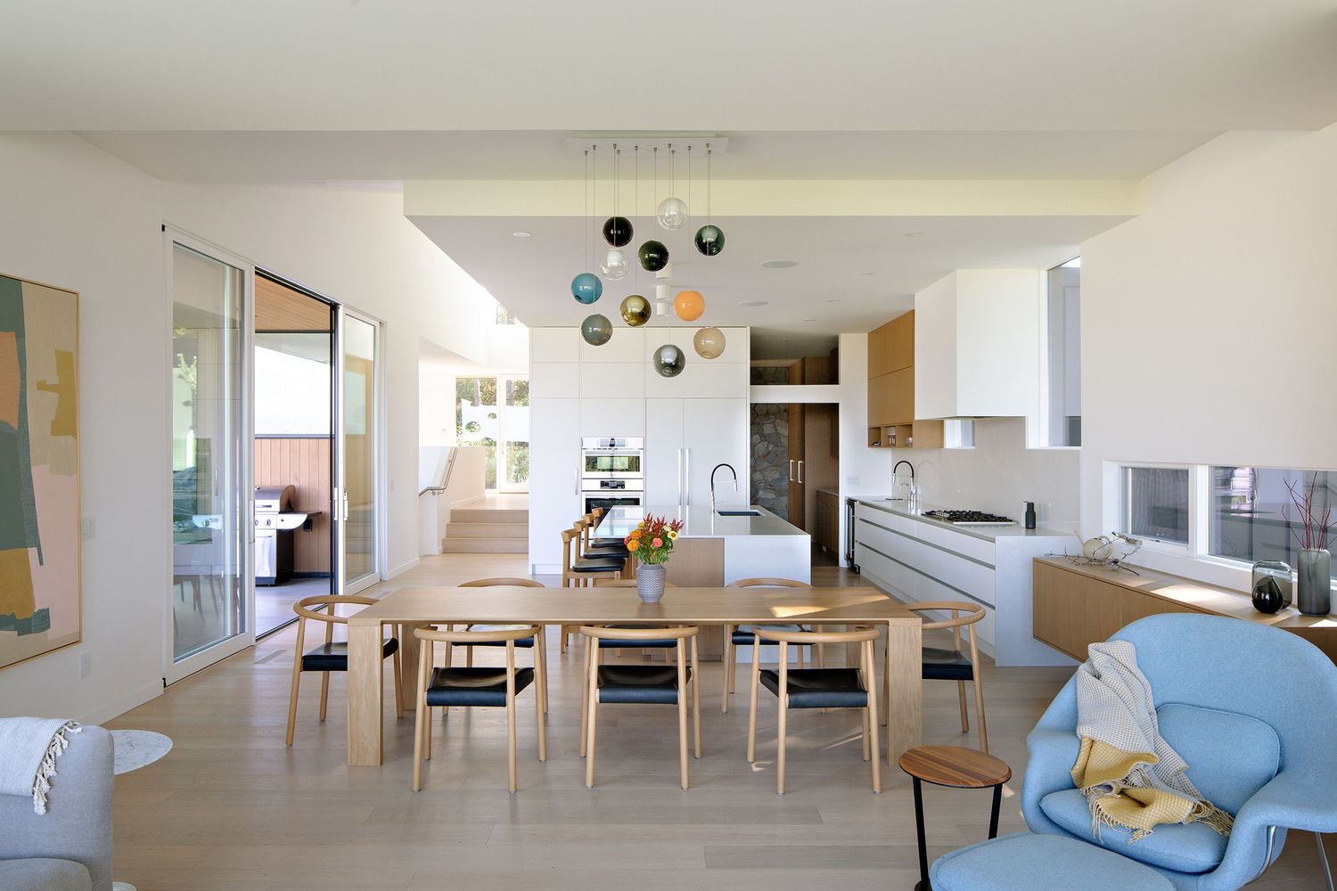 Modern open plan living space with light wood dining chairs, blue sofa, and colorful pendant lights hanging from ceiling.