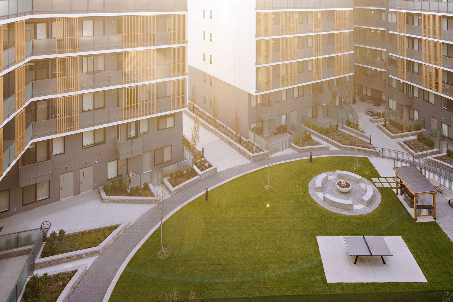 Curved modern apartment building surrounds a green courtyard with concrete paths and a circular fountain feature.