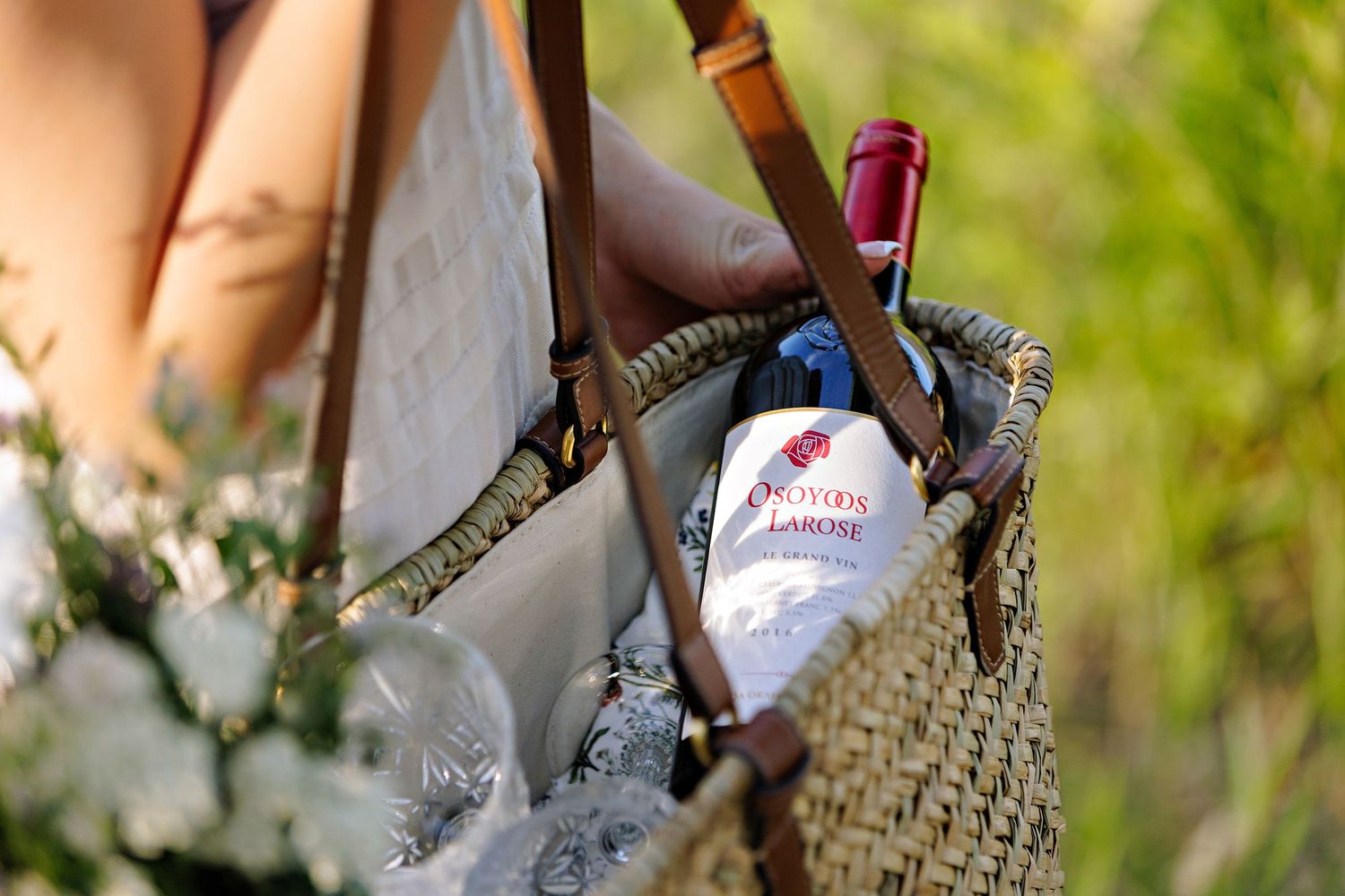 A bottle of wine peeking out of a woven straw beach bag with flowers in an outdoor setting.