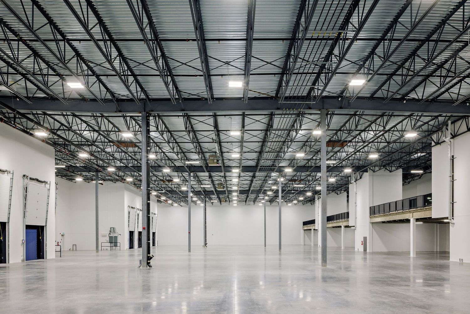 Large empty industrial warehouse with polished concrete floor, white walls, and metal ceiling structure with bright lighting fixtures.