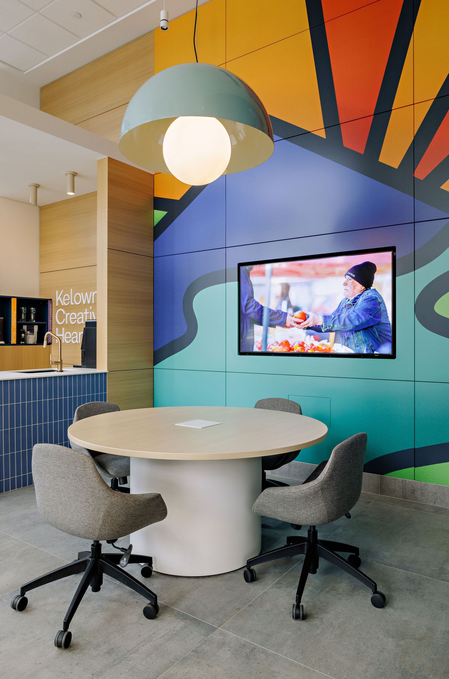 Modern office meeting space with round white table, grey chairs, and colorful geometric wall mural featuring mounted TV screen.
