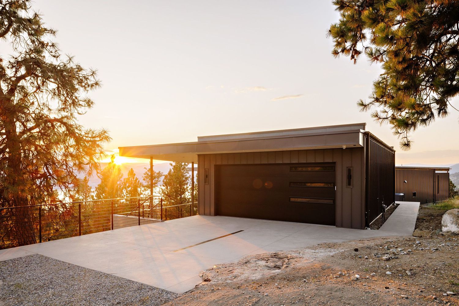 Modern garage with dark wood exterior and concrete driveway at sunset with golden light streaming through trees.
