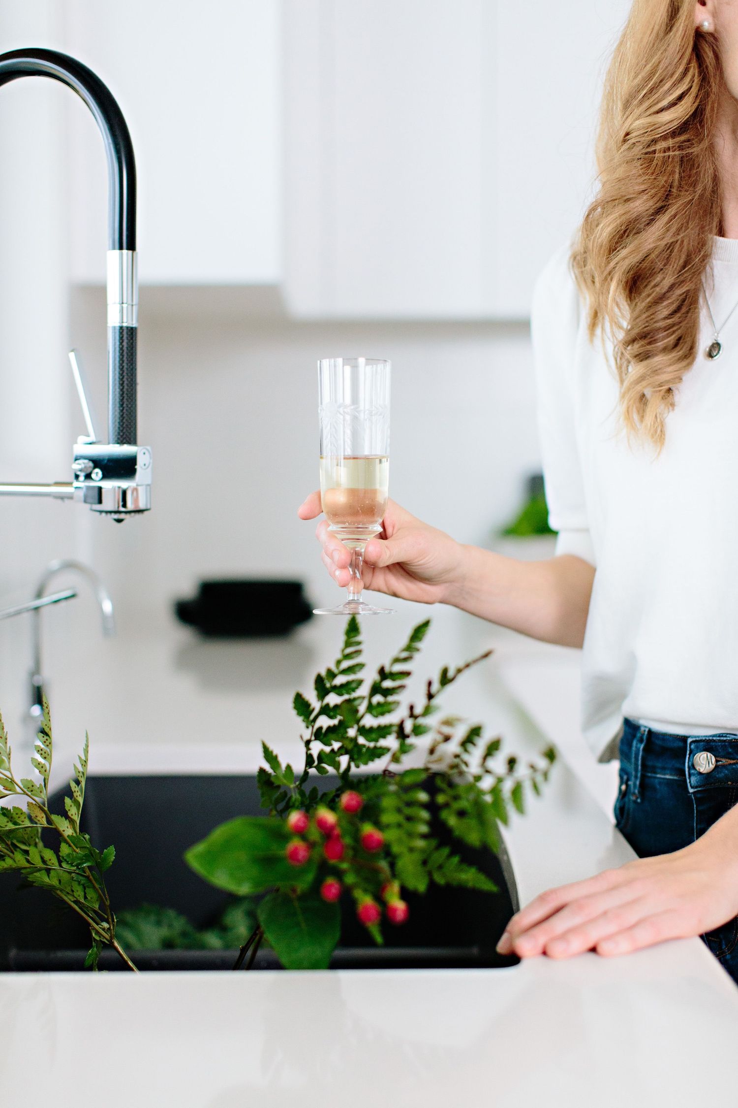 Hand holding a champagne glass near modern black kitchen faucet with greenery and berries on white counter.