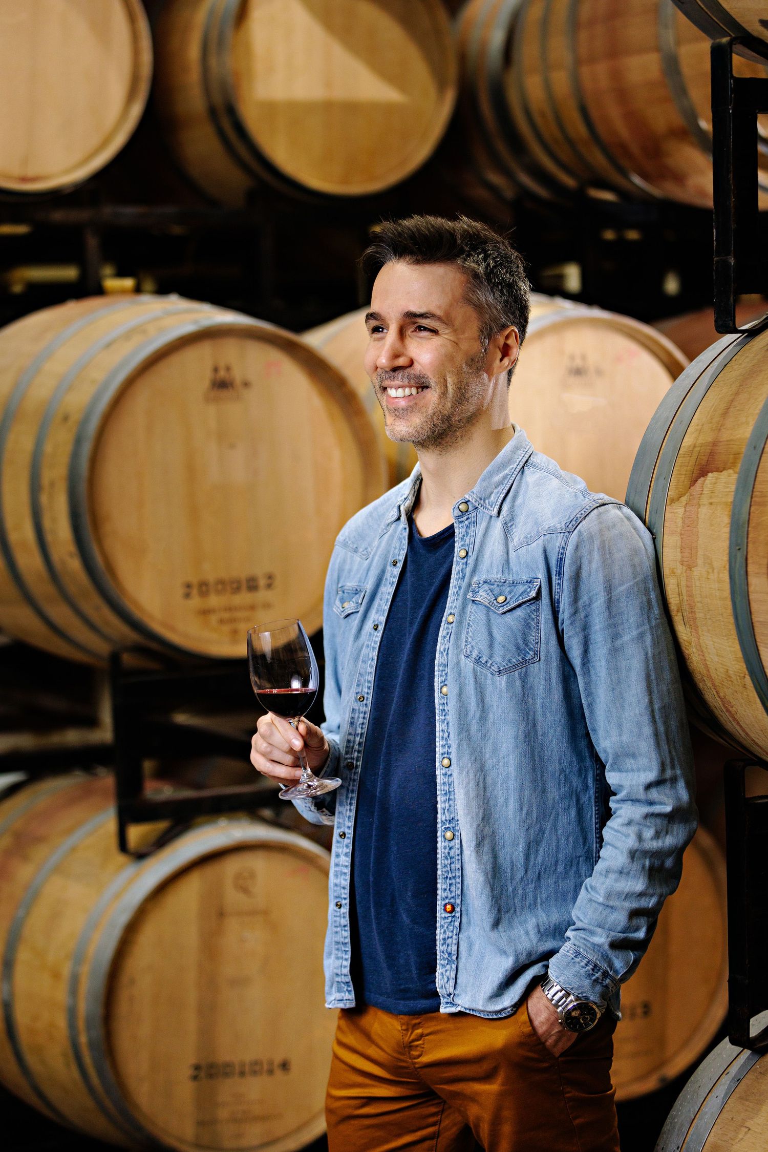 A person in denim shirt holding wine glass while standing next to oak barrels in a cellar winery.