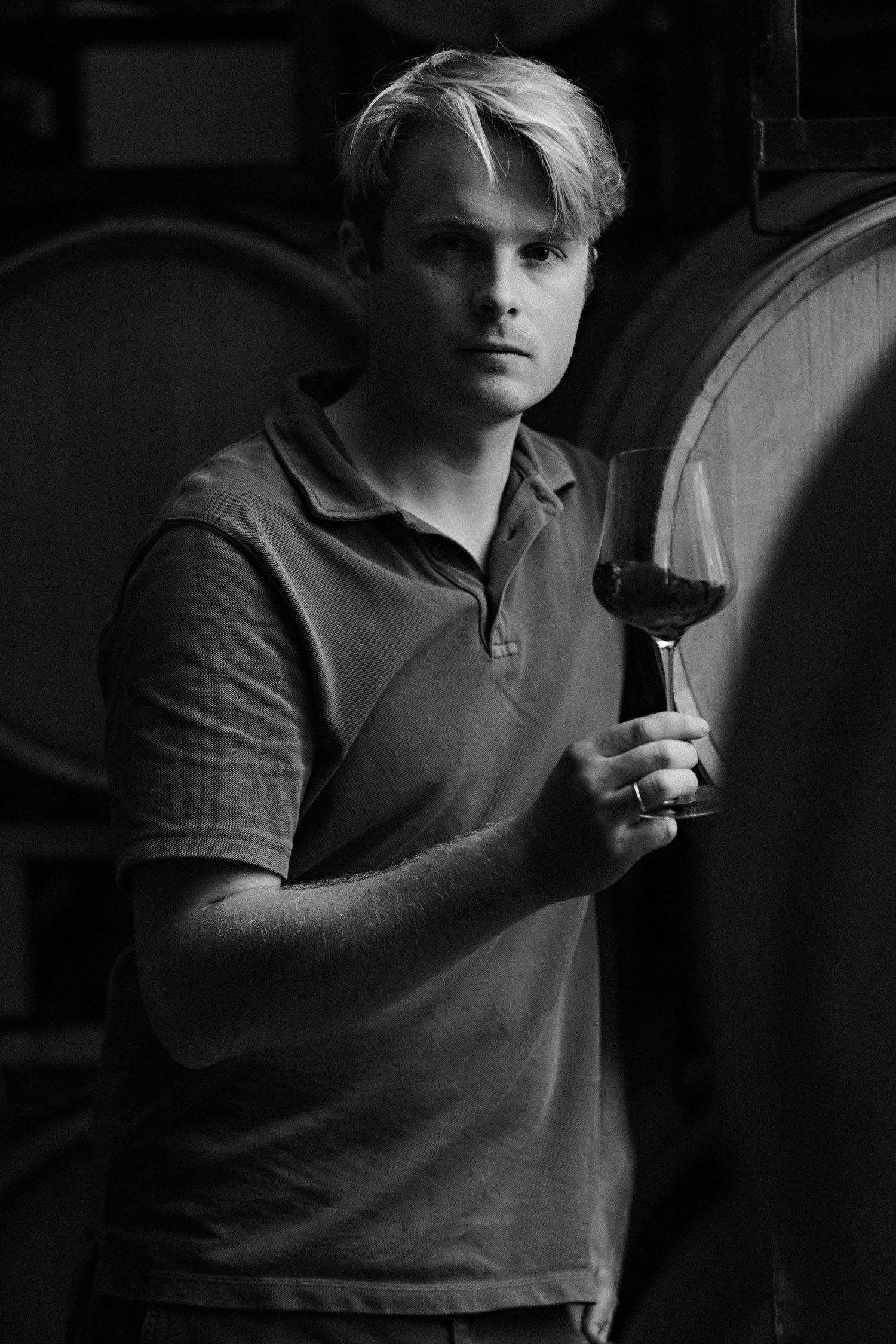 A sommelier in a polo shirt examines a glass of red wine in a dark cellar with wine barrels in the background.