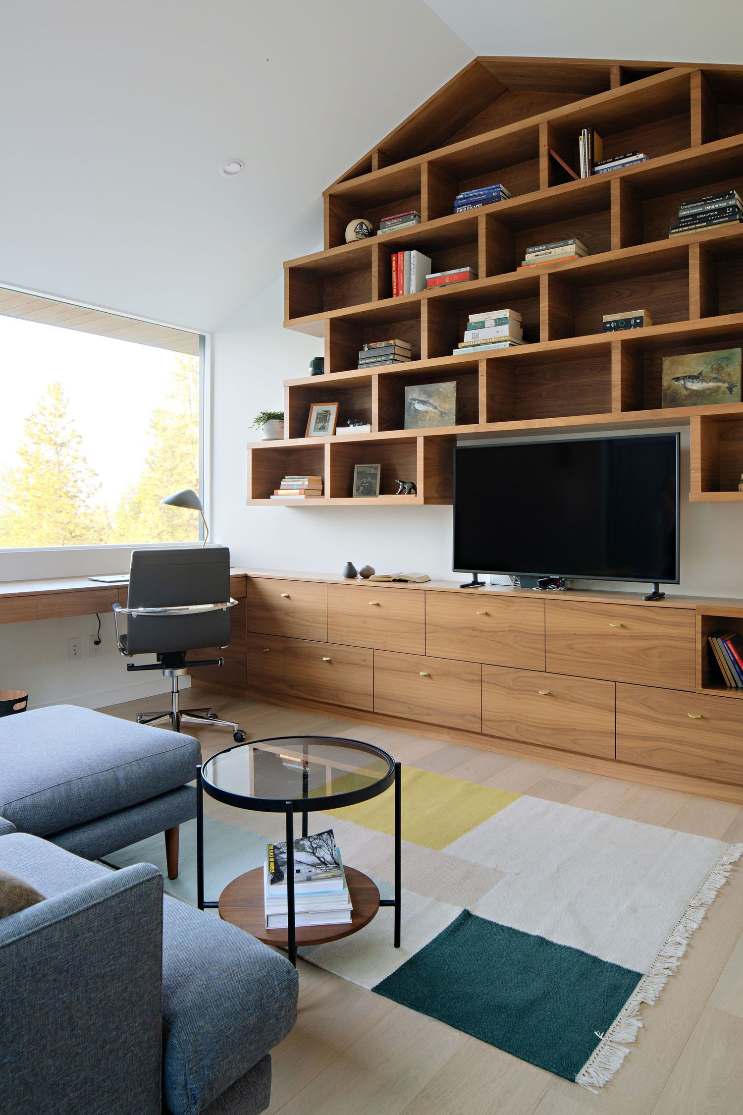 Modern living room with custom wood shelving unit, mounted TV, gray armchairs, and geometric patterned area rug.