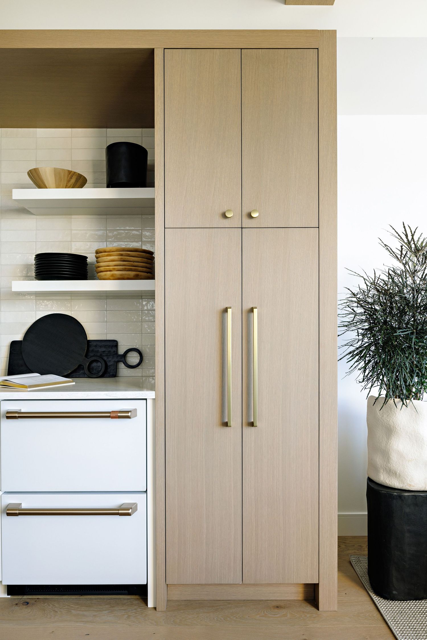 Modern light wood kitchen cabinet with brass handles next to white shelving unit displaying black cookware and dishes.