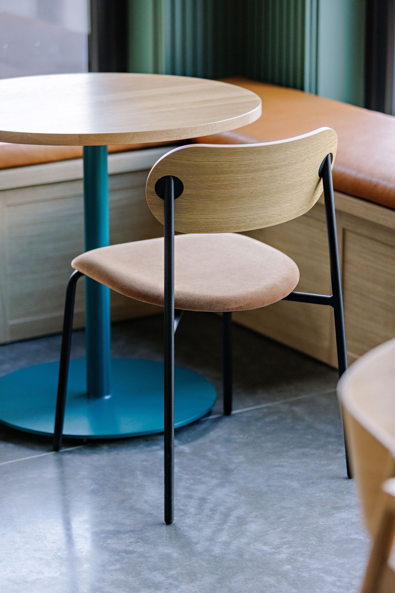 Modern minimalist dining chair with curved wooden backrest and beige cushioned seat next to round table on concrete floor.