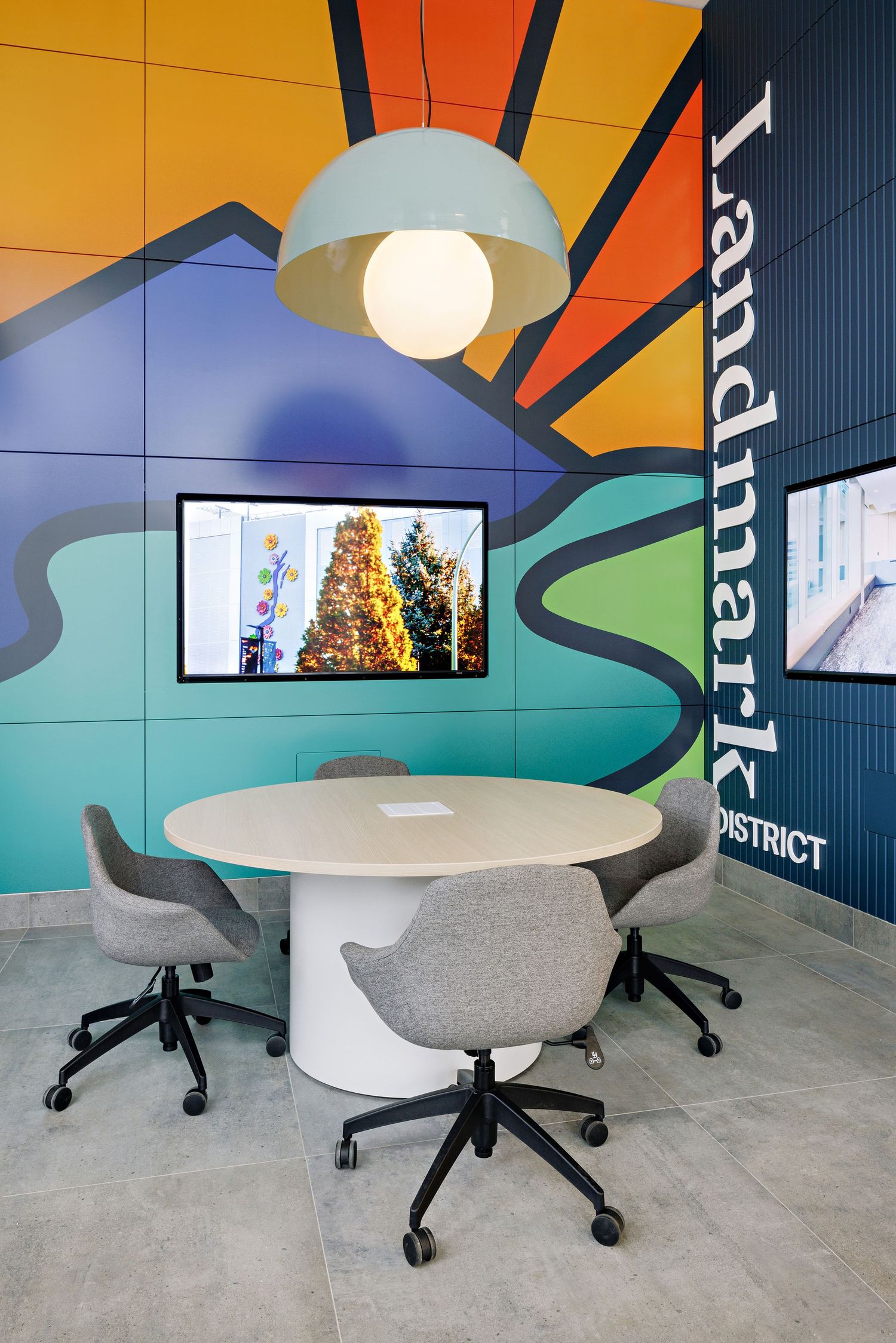 Modern meeting space with vibrant wall mural, round white table, grey chairs, and mounted TV screen showing holiday decor.