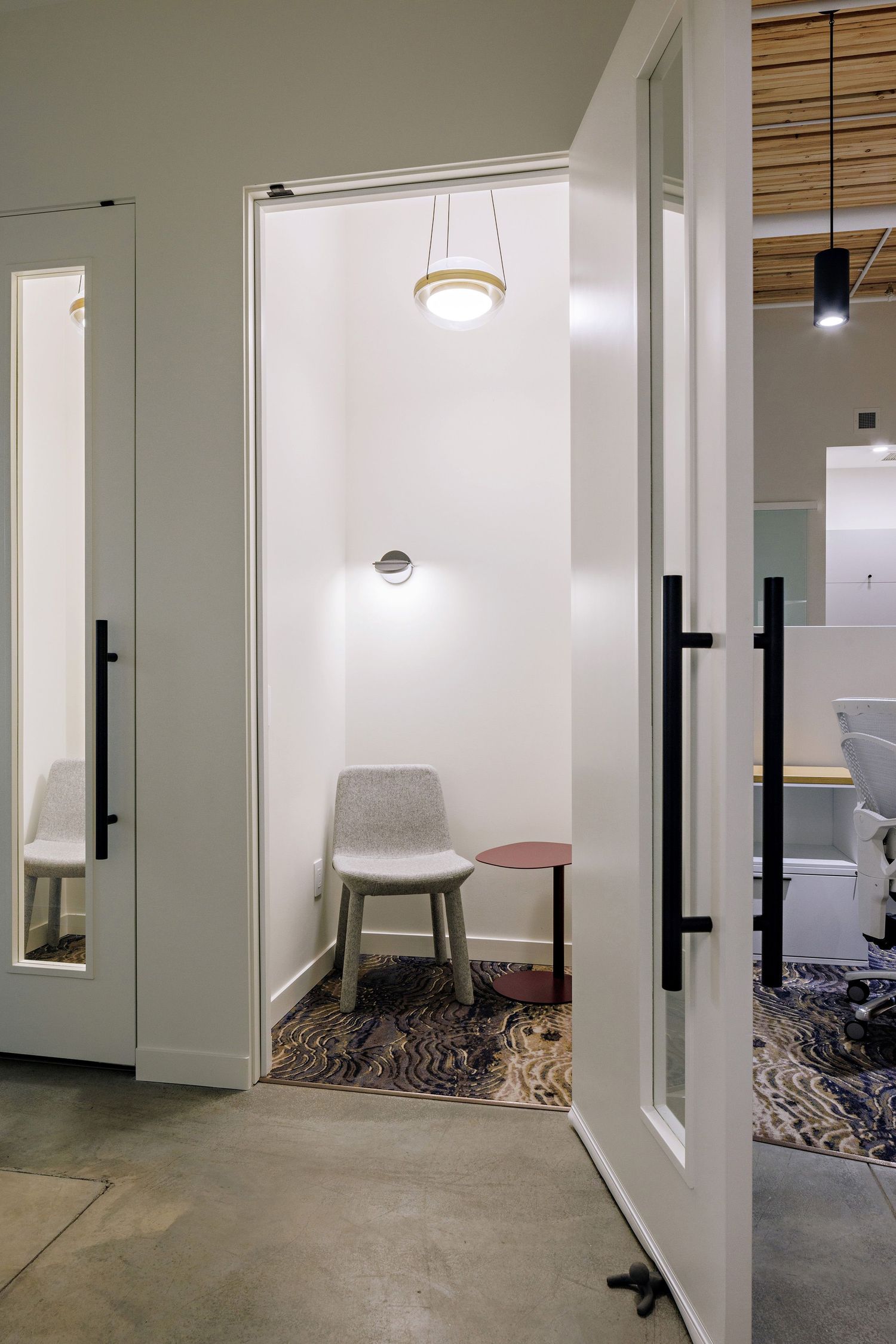 Small modern office phone booth with gray chair, pendant light, and glass doors in a minimalist workspace setting.