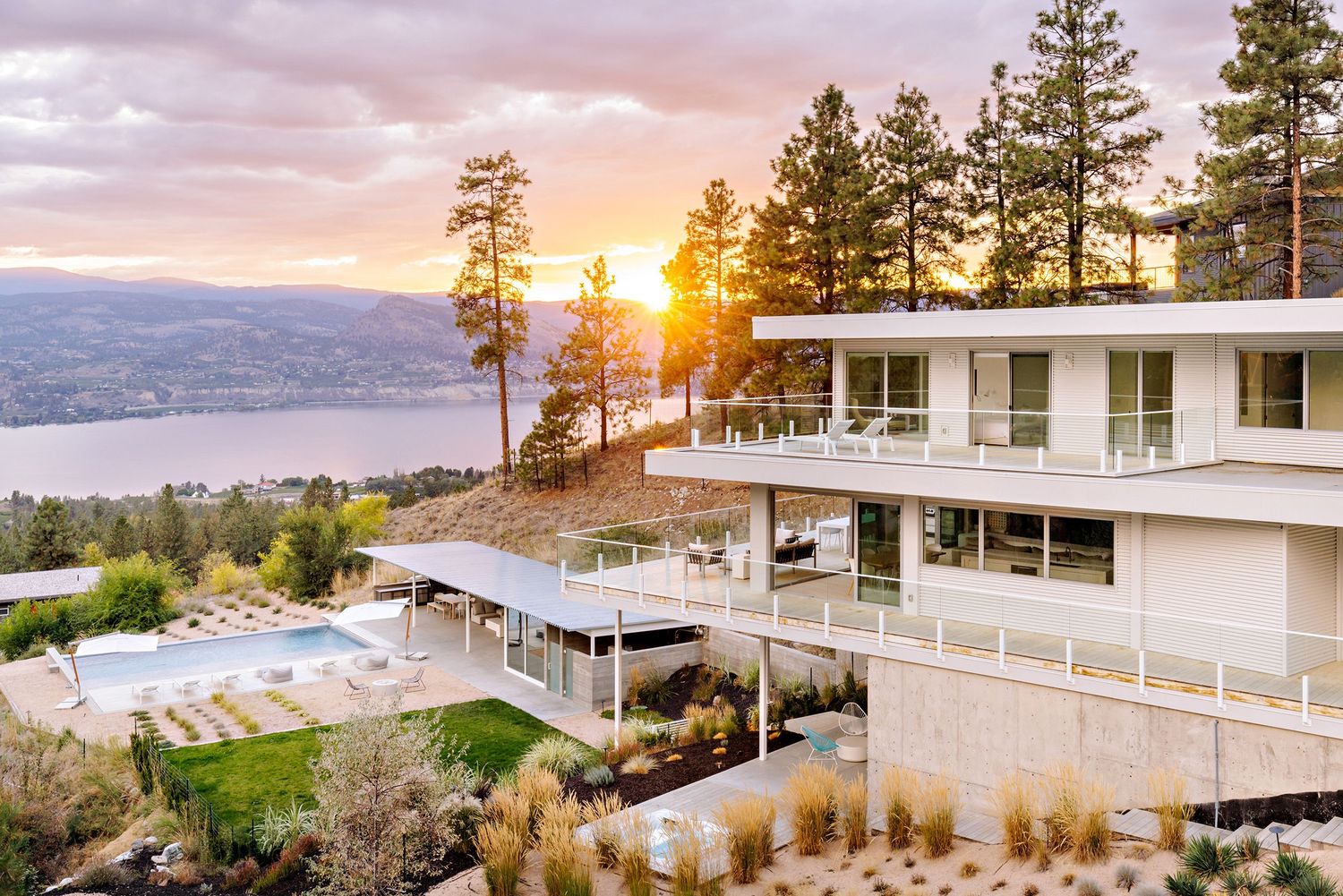 Modern luxury hotel with white exterior overlooking a lake at sunset with mountains and pine trees in the background.