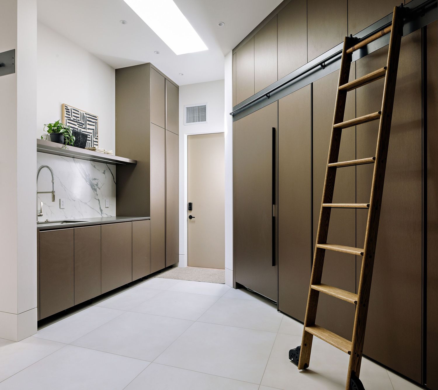 Modern minimalist kitchen with marble backsplash, brown cabinets and sliding library ladder against tall storage wall.
