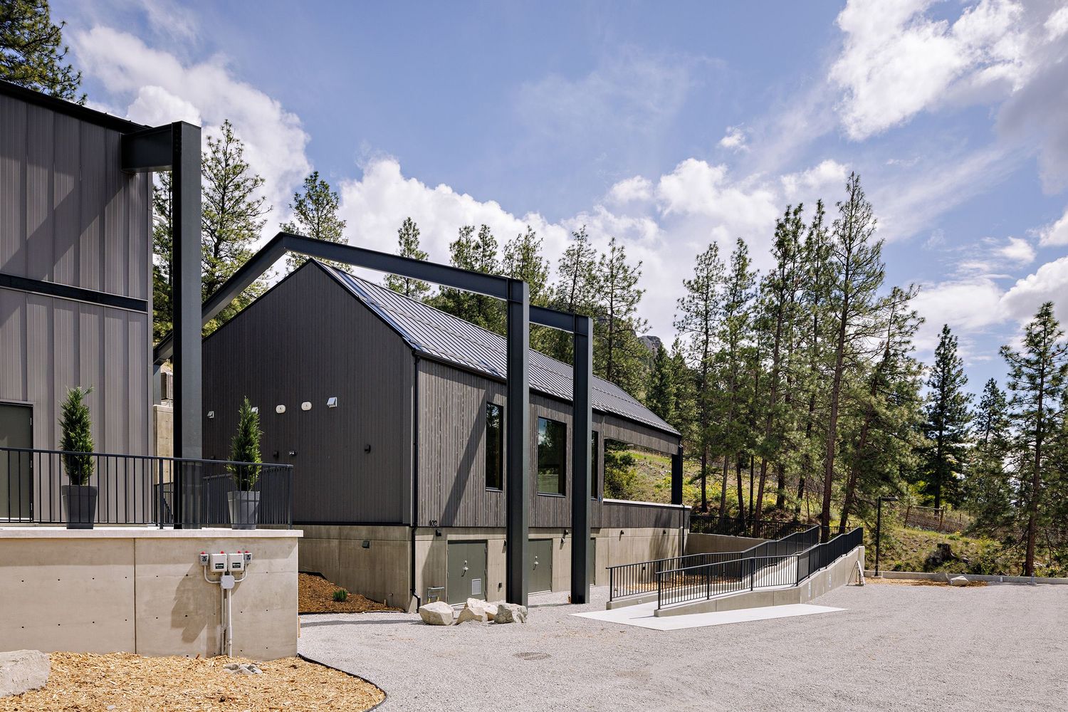 Modern black barn-style buildings with glass features nestled among pine trees under a bright blue sky with white clouds.