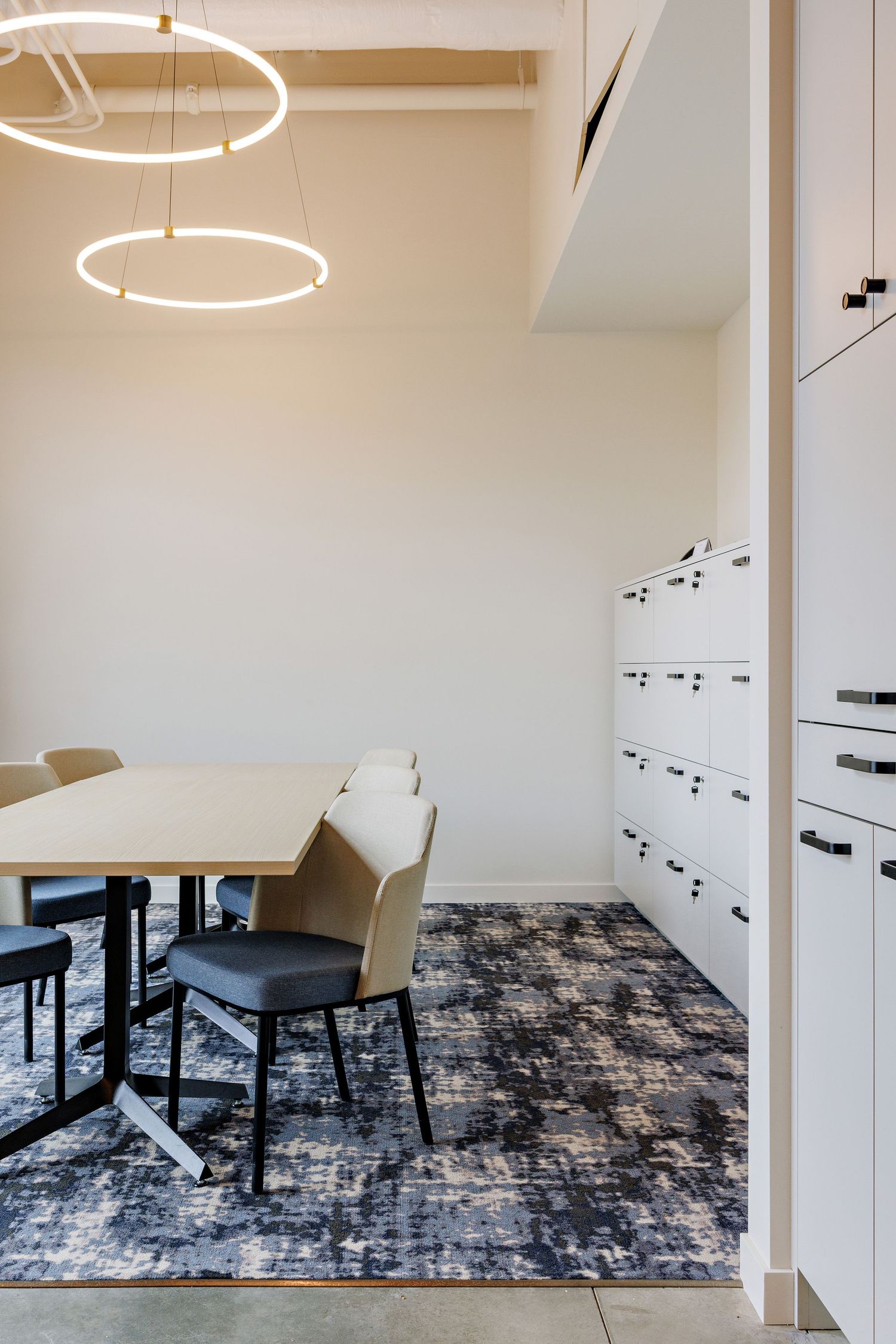 Modern office breakout space with circular pendant lights, round table, tan chairs, and built-in white storage lockers on a dark rug.