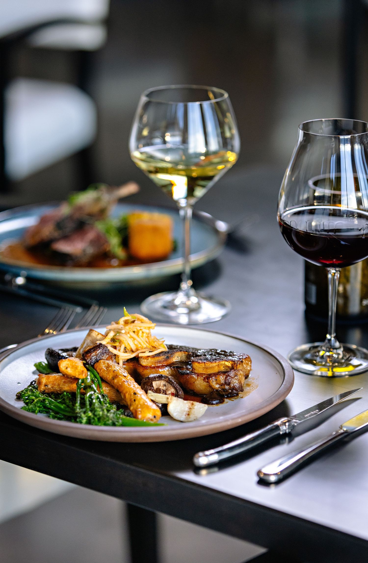 Fine dining plate with grilled meat, roasted vegetables and two wine glasses on a dark table setting.