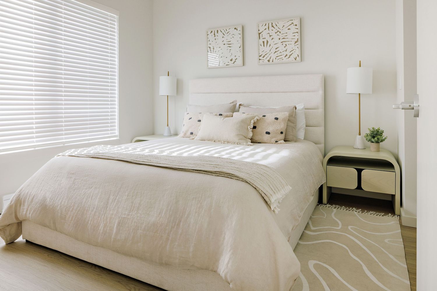 Modern minimalist bedroom with white bedding, neutral decor, and soft natural lighting through venetian blinds.
