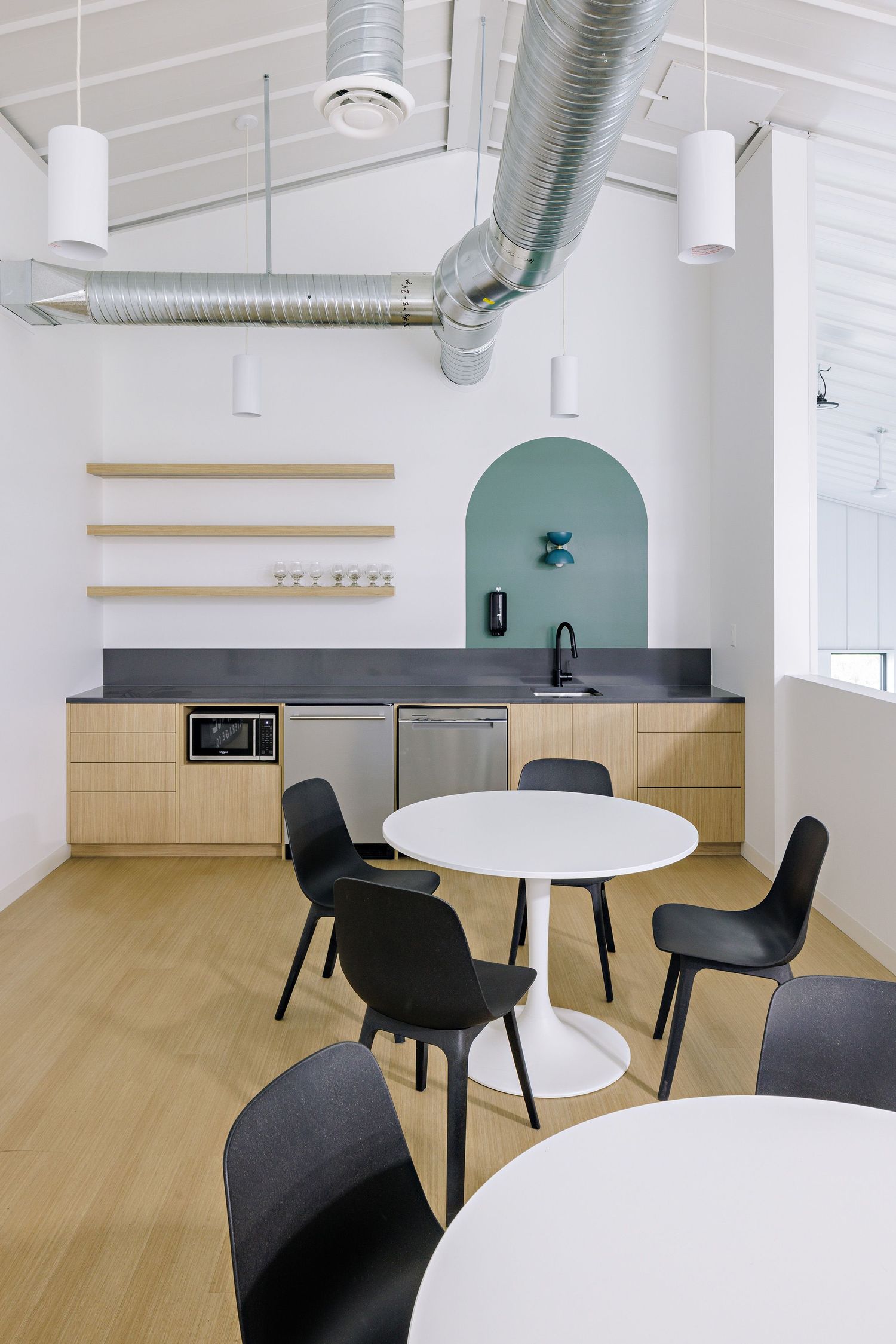 Modern office break room with white arched walls, wood cabinets, exposed ductwork, and round tables with black chairs.