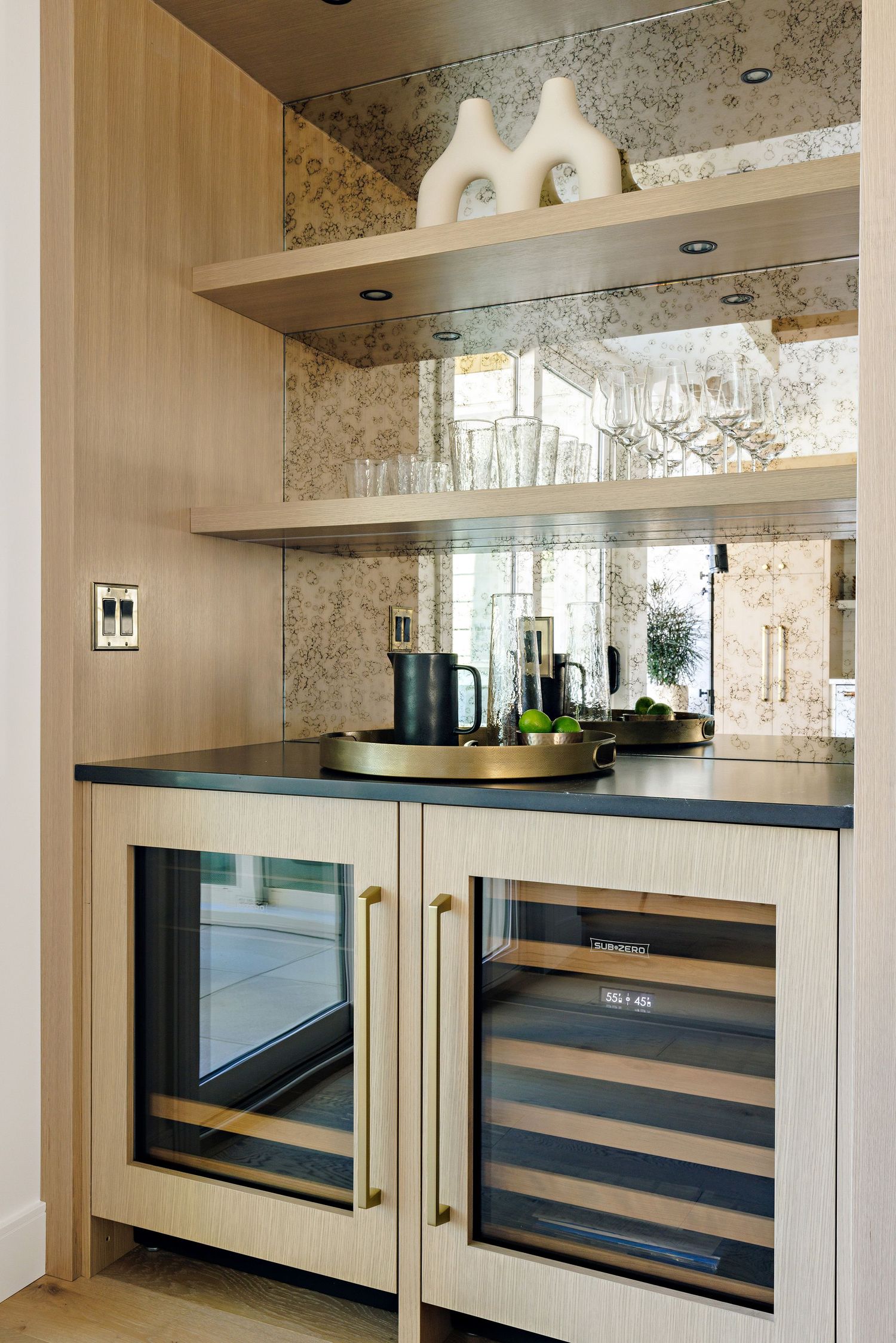 Modern built-in bar area with mirrored backsplash, wooden shelving, and dual wine refrigerators in light wood cabinetry.