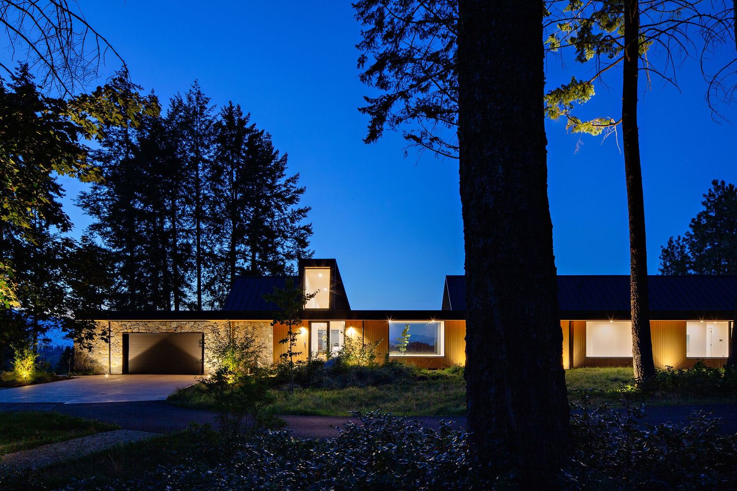 Modern single-story home illuminated at dusk with large windows and tall trees silhouetted against a deep blue sky.